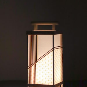May include: A rectangular wooden lantern with a warm, inviting glow. The lantern features a geometric design with vertical wooden slats and patterned paper panels. The top has a handle, and the base houses a small lightbulb. The overall design is minimalist and elegant.
