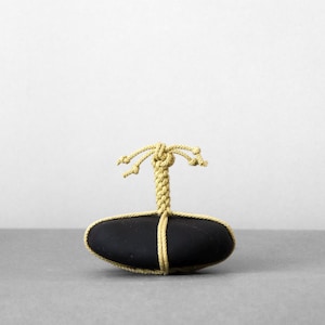 Puede incluir: Una piedra negra lisa de forma ovalada asegurada con un cordón decorativo de color amarillo claro. El cordón envuelve el centro de la piedra y se extiende hacia arriba en un asa vertical trenzada que termina en un nudo con cuatro extremos sueltos.
