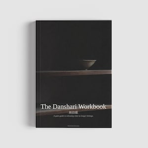 Könnte beinhalten: Ein dunkel gebundenes Buch mit dem Titel "The Danshari Workbook" mit japanischem Text und einer kleinen Schale auf einem Regal. Der Untertitel des Buches lautet: "Ein ruhiger Leitfaden, um loszulassen, was nicht mehr dazugehört." Der Text ist weiß.