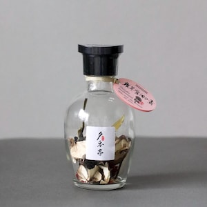 以下が含まれることがあります： 黒いキャップが付いた透明なガラス瓶で、乾燥したキノコとハーブが入っています。 日本語の文字が入った小さな白いラベルが瓶に貼られています。 日本語のテキストが入ったピンク色のタグが首からぶら下がっています。