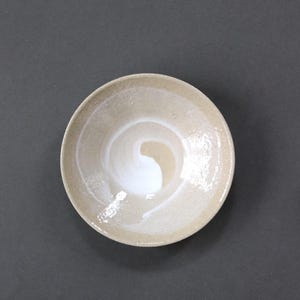 May include: A round ceramic bowl with a speckled beige and tan exterior and interior. The center of the bowl features a smooth, glossy white glaze applied in a swirling, spiral pattern that contrasts with the textured, matte finish of the clay.