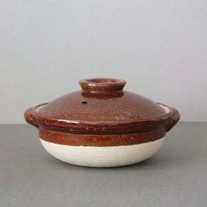 Puede incluir: Una olla de cocina de cerámica marrón con tapa a juego. La olla tiene una base blanca y una sección superior marrón con una tapa marrón oscura. La tapa tiene un asa pequeña y un orificio de ventilación.