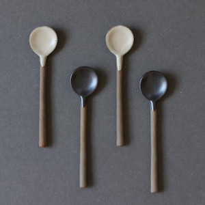 Puede incluir: Cuatro cucharas pequeñas con cabezas de cerámica y mangos de madera dispuestas sobre una superficie gris. Dos cucharas tienen cabezas blancas y dos tienen cabezas de color gris oscuro. Los mangos son de color marrón claro.