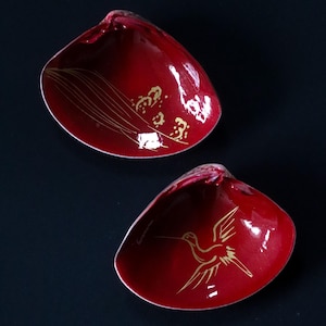 May include: Two deep red, shell-shaped dishes with a glossy finish. The top dish features delicate gold line art of tall grasses, while the bottom dish displays a gold illustration of a bird in flight. Both items have a smooth, curved interior.