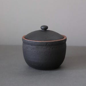 May include: A small, black ceramic jar with a lid. The jar has a rounded shape with a textured surface and a contrasting reddish-brown rim. The lid features a small, round knob.
