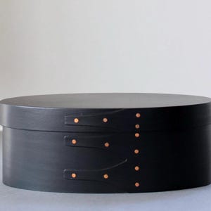 Puede incluir: Una caja negra de forma ovalada con tapa. La caja tiene tres correas de cuero con remaches de color cobre en la parte delantera y una fila de remaches en el lateral. La caja está sobre una superficie gris.