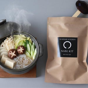 以下が含まれることがあります： 野菜、キノコ、豆腐が入った湯気の立つ日本の鍋が、木製の鍋敷きの上に置かれています。「Wabisabi Kitchen Nabe Kit Hot Pot Set」と書かれた茶色の紙袋が鍋の隣にあります。木のスプーンが袋の上に置かれています。