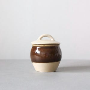 May include: A small, ceramic jar with a lid. The jar is a deep brown color in the middle, with a cream-colored base and lid. The lid has a small, arched handle. The jar is likely used for storage.
