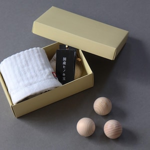 May include: A beige gift box with a white towel, a black tag with Japanese text, and three wooden spheres. The box is open, revealing the contents. The spheres are arranged on a gray surface.