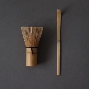 May include: A bamboo matcha whisk and scoop rest on a dark gray surface. The whisk has many thin tines bound together with a black string. The scoop is a long, flat piece of bamboo. This set is used for preparing matcha tea.