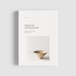 Op de afbeelding: Een wit hardcover boek met de titel WHAT WE ACTUALLY EAT: A field guide to the everyday Japanese kitchen. De kaft bevat een gecentreerde foto van een enkele, lichtgekleurde, conische keramische kom tegen een effen, neutrale achtergrond.