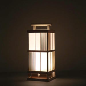 May include: A rectangular table lamp with a light wood frame and a handle on top. The lamp features panels of translucent white material alternating with sections of vertical wooden slats. A warm light glows from within the structure.