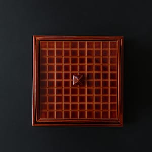 May include: Square, brown ceramic tile tray with a grid pattern and a raised, geometric center. The tray has a dark brown frame and is set against a black background.