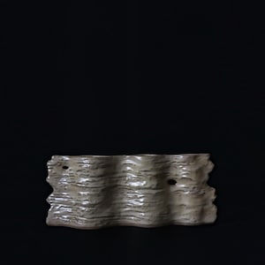 Puede incluir: Un florero de cerámica beige con una silueta horizontal ondulada y un acabado de superficie texturizado similar a la corteza. El objeto presenta tres aberturas cilíndricas distintas en el borde superior y dos pequeñas perforaciones circulares.
