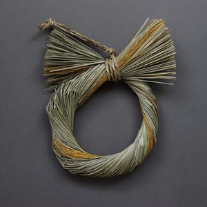以下が含まれることがあります： 天然の乾燥草で作られた手作りのリース。円形デザインで、上部にリボンのような装飾があります。リースは主に緑と茶色で、金色のアクセントがあり、家の装飾に適しています。