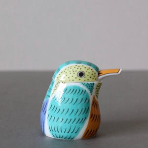 May include: A ceramic bird-shaped container with a lid. The bird has a turquoise body with blue and orange accents. The head is yellow with black dots and a long orange beak. The container is likely for decorative or functional use.