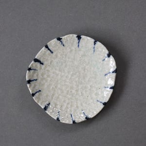 以下が含まれることがあります： テクスチャ加工された表面と不規則な丸みを帯びた形状の、小さなオフホワイトのセラミックプレート。プレートは、縁に沿って濃い青色の垂直な筆致で装飾されています。手作りのこの皿は、少量の料理の提供に最適です。