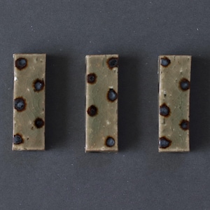 Puede incluir: Tres baldosas cerámicas rectangulares con un diseño moteado. Las baldosas son de color verde apagado con manchas azul oscuro y marrón. Las baldosas están dispuestas verticalmente sobre un fondo gris oscuro.