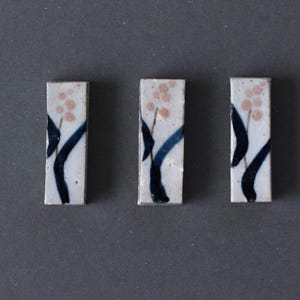 May include: Three rectangular ceramic chopstick rests. Each rest has a white background with a floral design. The design includes pink flower clusters, dark blue stems, and black leaves. The chopstick rests are arranged on a grey surface.