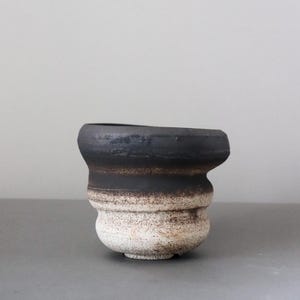Puede incluir: Una taza de cerámica con un borde ancho y acampanado y un cuerpo redondeado. La parte superior presenta un acabado negro mate, mientras que la sección inferior transiciona a una superficie texturizada moteada en blanco roto y marrón claro.