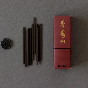 Pode incluir: Uma caixa vermelha escura com texto dourado, um conjunto de varetas de incenso castanho escuro e um pequeno objeto redondo e escuro. A caixa é retangular e o texto está em língua estrangeira. Sobre uma superfície cinzenta.
