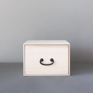 May include: A small, rectangular wooden storage box with a light, natural wood grain finish. The front features a single drawer equipped with a curved, black metal pull handle centered on the face. The box has a clean, boxy frame design.