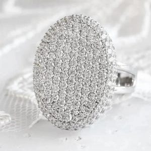 Könnte beinhalten: Ein ovaler Ring, besetzt mit vielen kleinen, glitzernden Steinen. Der Ring hat ein silbernes Band, und die Steine erzeugen eine helle, auffällige Darstellung. Der Ring ist vor einem weichen, weißen Hintergrund platziert. Es ist ein Schmuckstück.