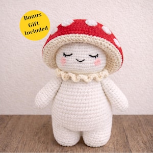 May include: A crocheted mushroom doll with a red and white spotted cap, closed eyes, and rosy cheeks. The doll has a cream-coloured frilly collar and is made of white yarn. A yellow circle with the text "Bonus Gift Included" is in the upper left corner.