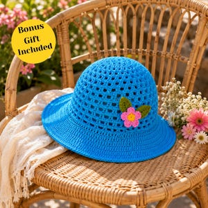 Può includere: Un cappello da sole all'uncinetto blu brillante con una tesa larga, decorato con un fiore rosa e giallo e foglie verdi. Il cappello è esposto su una sedia di vimini. Il testo "Bonus Gift Included" è in un cerchio giallo.