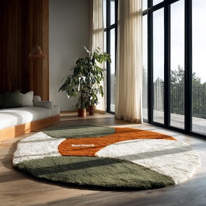 以下が含まれることがあります： モダンなデザインの丸いシャギーラグ。ラグは緑、オレンジ、白のセクションで構成されています。「Bloom Haven IN」の文字がラグにプリントされています。明るい部屋の木製の床にラグが置かれています。