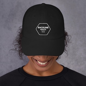 Puede incluir: Gorra de béisbol negra con un logotipo hexagonal blanco que dice "BACKLINE SUPPLY CO. EST. 2019". La gorra está hecha de un material suave y tiene una visera curva. La gorra la lleva una persona.