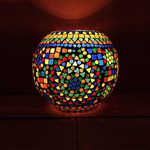 Puede incluir: Un portavelas redondo de cristal de mosaico colorido. El portavelas está iluminado desde dentro, mostrando un vibrante patrón de formas geométricas rojas, naranjas, amarillas, verdes, azules y moradas. El portavelas está sobre una superficie de madera oscura.