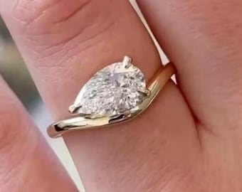 May include: A gold ring featuring a pear-shaped diamond. The diamond is set in a delicate, swirling band. The ring is shown on a person's finger, highlighting the elegant design and sparkle of the gemstone.