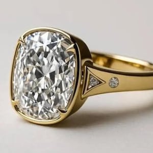May include: A gold ring featuring a large, cushion-cut diamond. The diamond is set in a bezel setting, and the band has a simple, elegant design with small accent diamonds. The ring is displayed on a white surface.