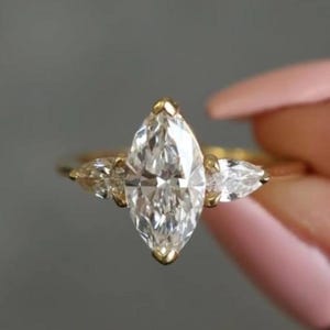 May include: A gold ring with a large, marquise-cut diamond as the central stone, accompanied by two smaller pear-shaped diamonds. The ring's band is slender, set against a blurred backdrop.