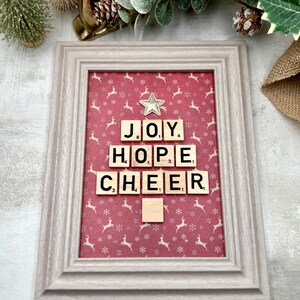 May include: A framed Christmas decoration with a red background patterned with white reindeer and snowflakes. Wooden letter tiles spell out "JOY, HOPE, CHEER" in a Christmas tree shape. A star embellishment is above "JOY". The frame is light grey.