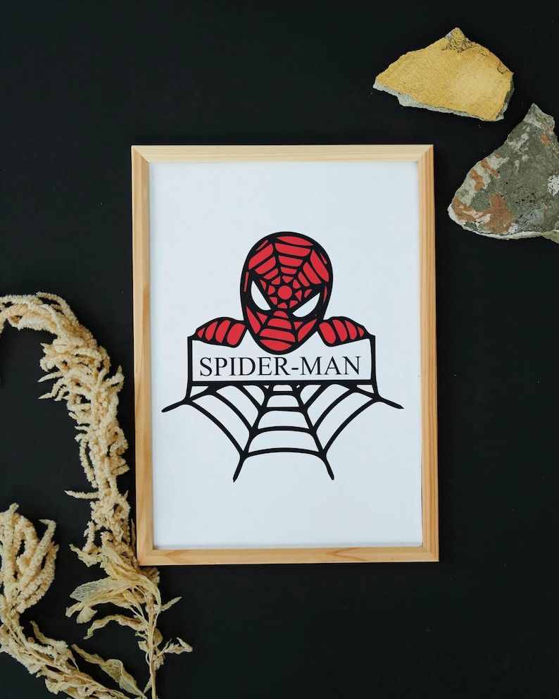 May include: Framed artwork featuring a red and black Spider-Man design. The image shows Spider-Man's head and a web, with the text "SPIDER-MAN" below. The artwork is in a light wood frame, set against a black background.