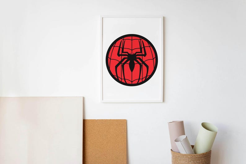 May include: Framed artwork featuring a black spider silhouette on a red webbed sphere. The artwork is displayed in a white frame against a white wall. The image evokes a comic book or superhero theme.