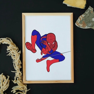 May include: Framed artwork featuring a stylized illustration of Spider-Man in a dynamic pose. The superhero is depicted in his classic red and blue costume, with a web extending from his hand. The artwork is set against a white background within a natural wood frame.