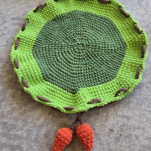 May include: A handmade, round crochet rug in shades of green with brown stitching around the edge. Two orange carrot-shaped embellishments hang from the bottom. The rug has a textured, spiral design.