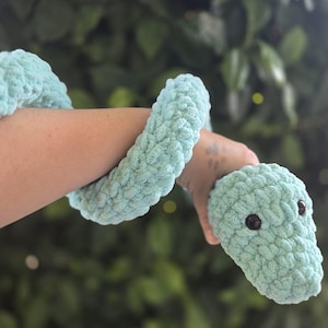 Puede incluir: Un juguete de serpiente de ganchillo azul claro con ojos marrones oscuros. La serpiente de peluche está envuelta alrededor de un brazo, con la cabeza de la serpiente sostenida en una mano. La serpiente está hecha de un hilo suave y texturizado.
