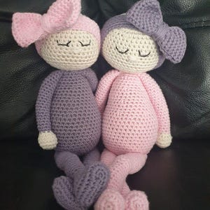 May include: Two handmade crochet dolls with round cream-coloured heads, closed eyes, and small noses. One doll wears a purple body with a pink bow, while the other wears a pink body with a purple bow. Both dolls have matching cream-coloured hands and feet.