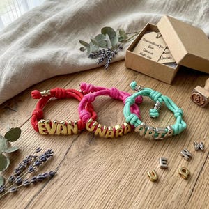 Könnte beinhalten: Drei personalisierte Armbänder in Rot, Pink und Türkis. Jedes Armband hat einen Namen, der mit goldenen oder silbernen Buchstabenperlen geschrieben ist. Die Armbänder werden auf einer Holzoberfläche mit einer Geschenkbox und dekorativen Elementen präsentiert.
