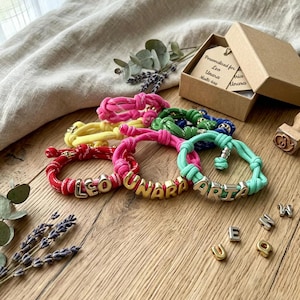 May include: A collection of colorful, knotted bracelets with gold and silver letter beads spelling out names. The bracelets are in shades of red, pink, yellow, green, and blue. A small gift box and loose letters are also visible.