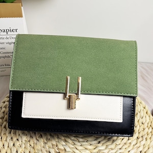 Pode incluir: Uma bolsa com blocos de cores com aba verde com efeito camurça, painel central branco e base preta. A bolsa apresenta um fecho dourado e formato retangular, adequada para uso diário.