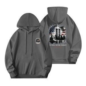 Puede incluir: Una sudadera con capucha gris carbón que presenta un pequeño gráfico en el pecho con la Estatua de la Libertad y el texto NEVER FORGET. La parte posterior muestra un gráfico grande de las Torres Gemelas, la Estatua de la Libertad y la bandera.