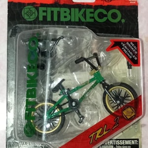 May include: A green and black Fitbikeco TRL 2 model BMX bike in a plastic package. The packaging includes the Flick Trix logo and the words "Real Brakes". The bike has gold-coloured rims and black tyres. The packaging also includes a warning about small parts.
