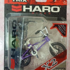 May include: A miniature purple and white Haro Bikes X2 BMX bike toy in a clear plastic package. The packaging features the Flick Trix logo and the words "Real Brakes" and "Vératables Freins". The toy is recommended for ages 9 and up.