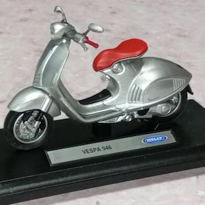 May include: A silver Vespa 946 model scooter with a red seat and black tyres. The miniature scooter is displayed on a black base with the brand name "Welly" visible. The scooter has a detailed design.