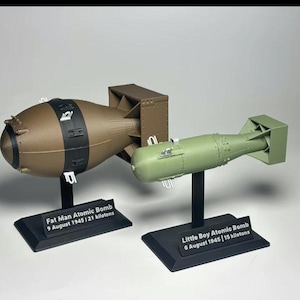Peut inclure: Deux modèles de bombes atomiques, "Fat Man" et "Little Boy", sont présentés. "Fat Man" est marron avec des bandes noires, et "Little Boy" est vert. Chacun repose sur un socle noir avec le texte : "Fat Man Atomic Bomb 9 August 1945 | 21 kilotons" et "Little Boy Atomic Bomb 6 August 1945 | 15 kilotons".
